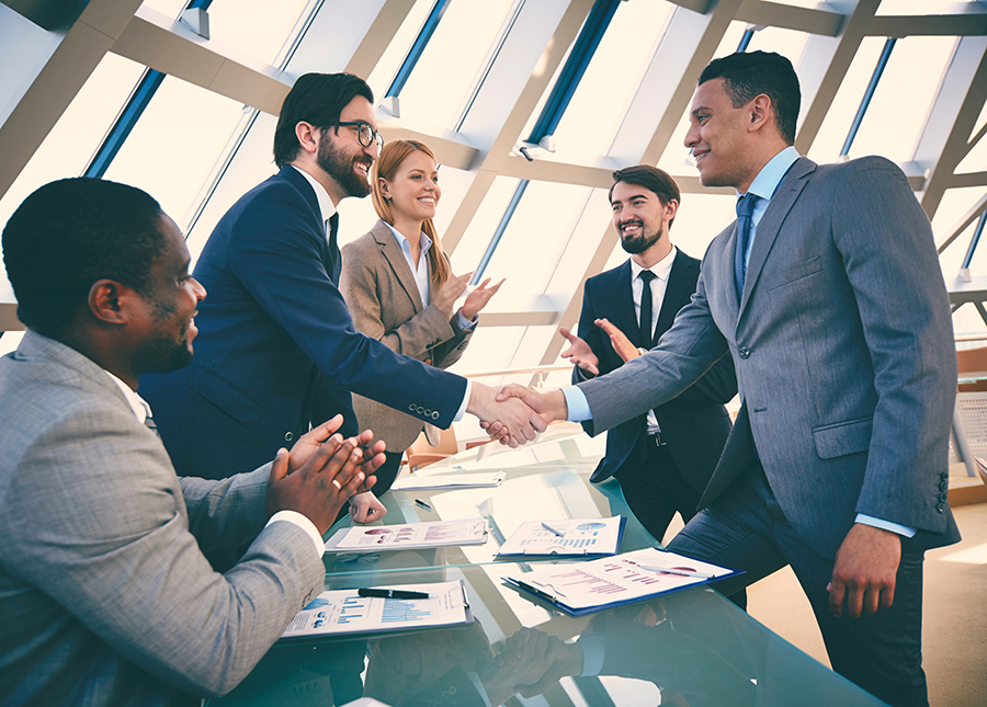 Business professionals shaking hands with colleagues applauding in a modern office.