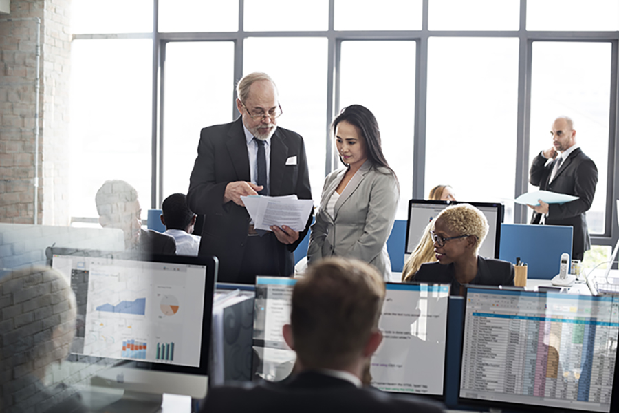 Senior manager and employee reviewing documents in a busy corporate office environment.