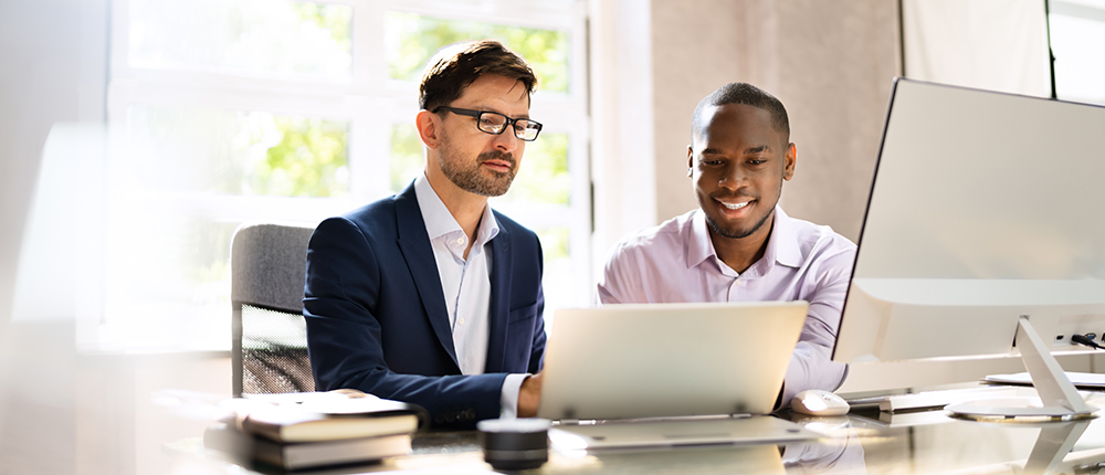 Two professionals collaborating on a project with laptops in a bright office setting.