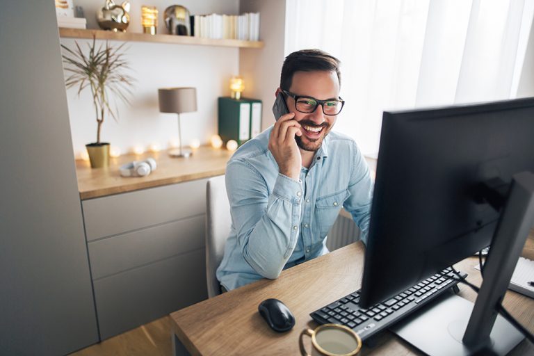 Smiling man in glasses on phone call with computer at home office.