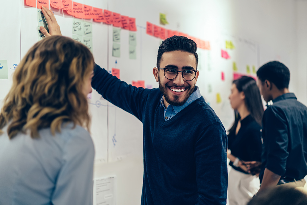 Smiling man in glasses at a collaborative workspace with sticky notes on the wall.