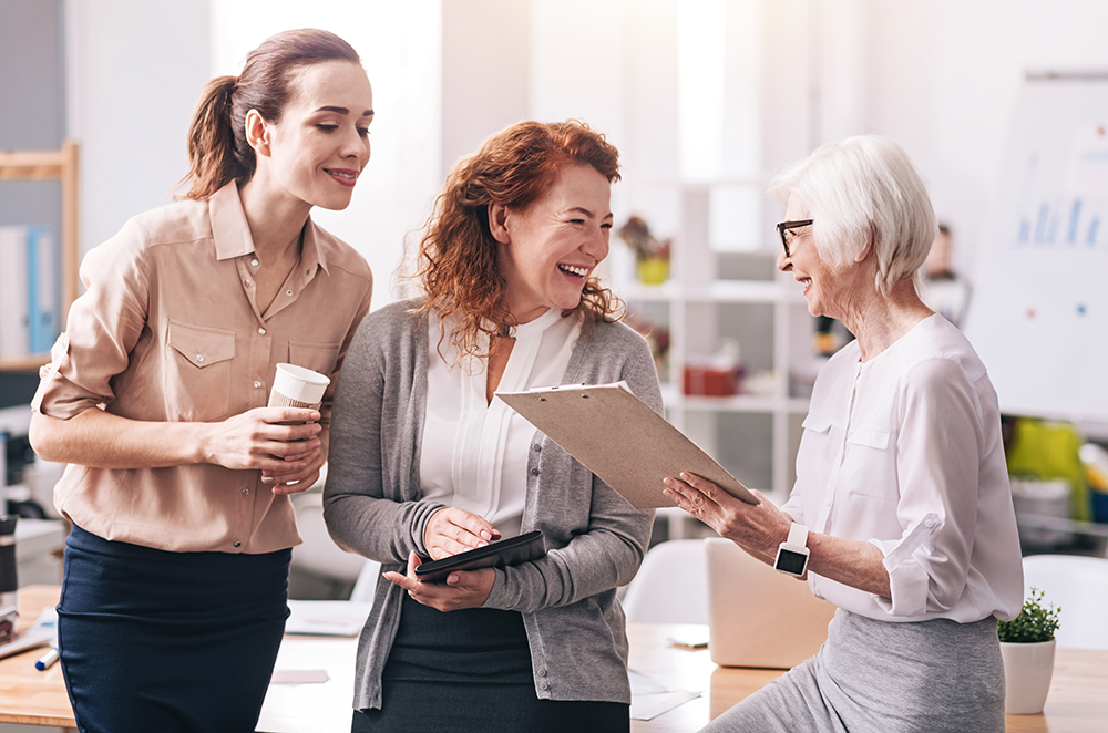 Three professional women of varying ages smiling and discussing over a clipboard in an office.