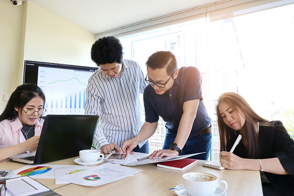 Team of four professionals analyzing data on computer and printed graphs in sunlit office.