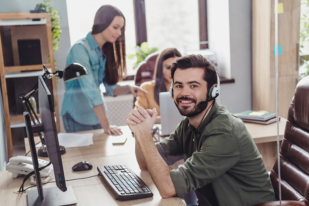 Cheerful male customer service representative with headset at computer in busy office.