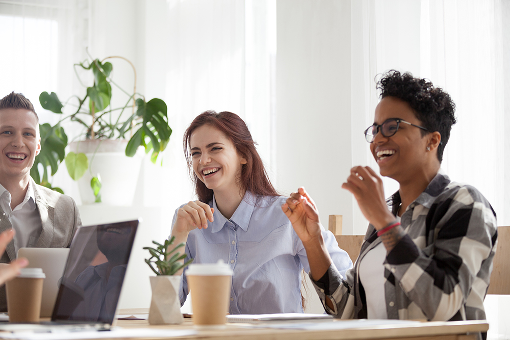 Three diverse colleagues laughing and enjoying a teamwork session with laptops and coffee.