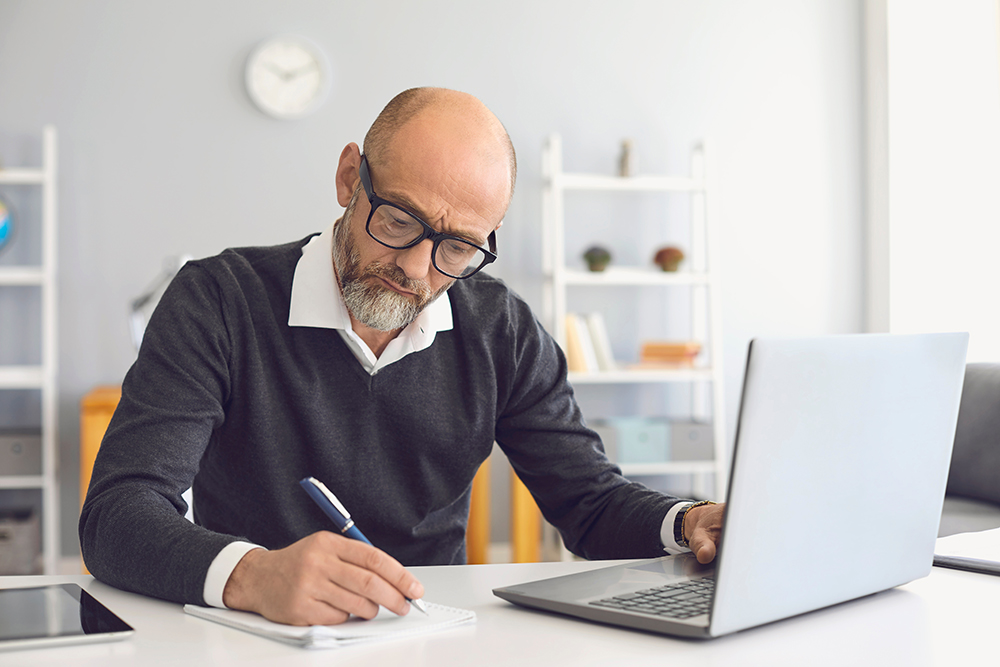 Senior man with glasses writing notes while using a laptop in a modern office.