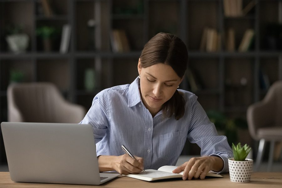 Professional woman concentrating on writing notes with a laptop on desk.