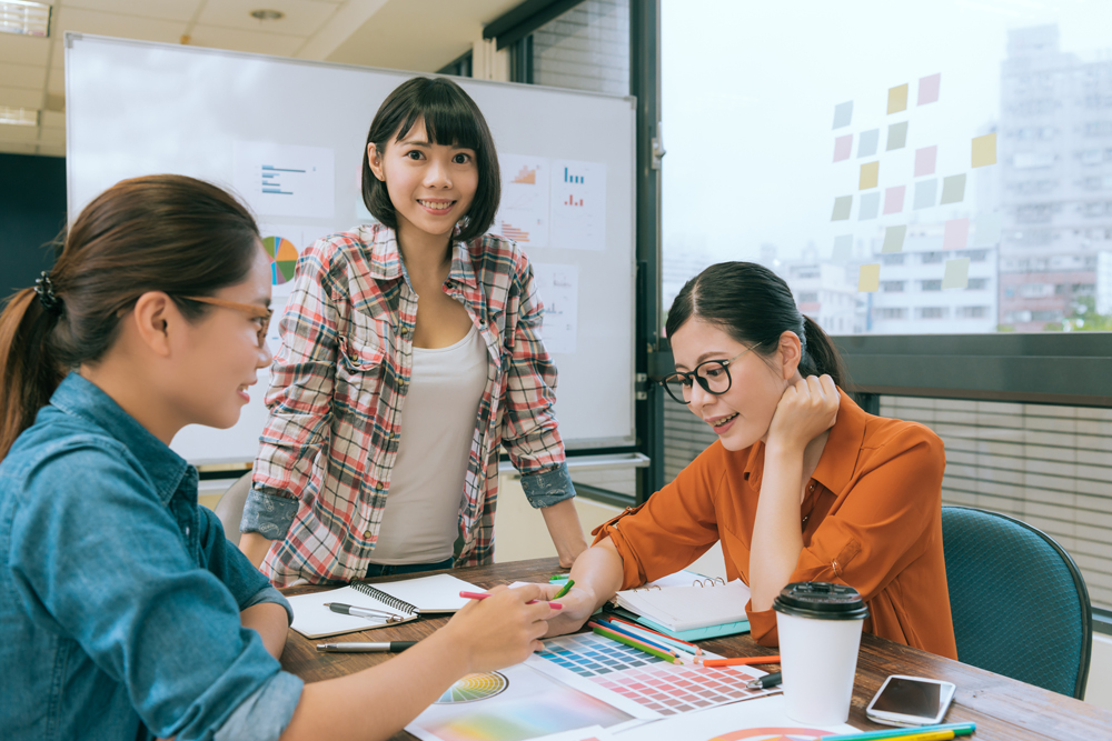 Three female colleagues discussing project details with color swatches on desk in bright office.