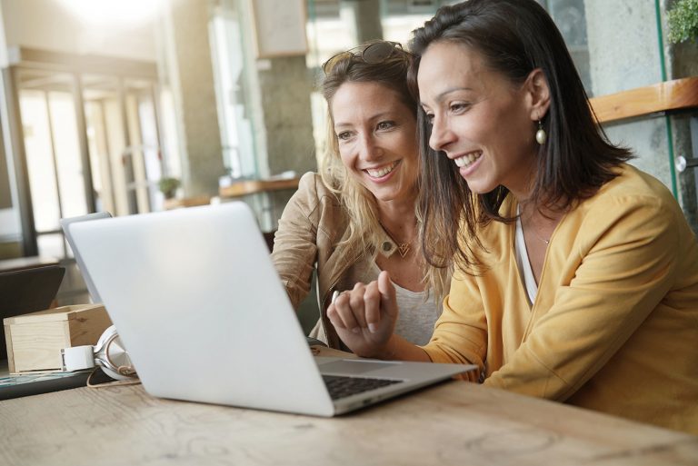 Two cheerful women collaboratively working on a laptop at a casual cafe setting.