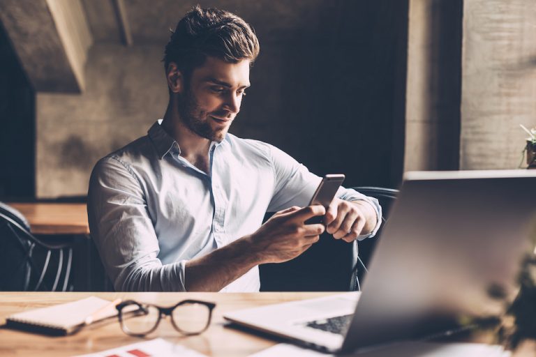 Handsome young businessman using smartphone while working at a rustic office desk with a laptop.