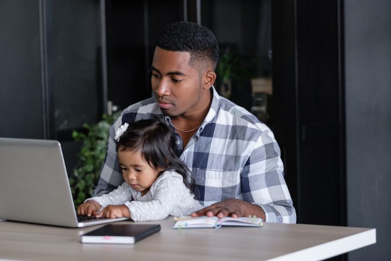 Focused father working on laptop while his young daughter plays beside him at home office desk.