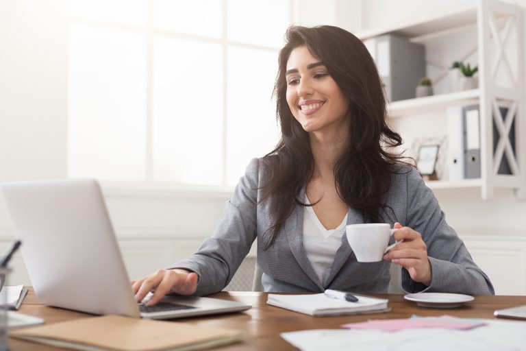 Cheerful businesswoman with coffee using a laptop in a well-lit home office environment.