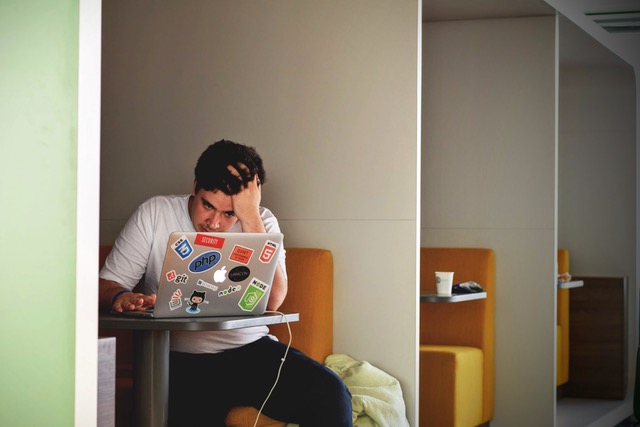 Stressed young man with his hand on his head, looking at a laptop covered in stickers at a cafe.