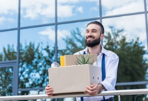 Smiling man carrying a box of personal belongings, standing outside an office building, symbolizing leaving a job or resigning.
