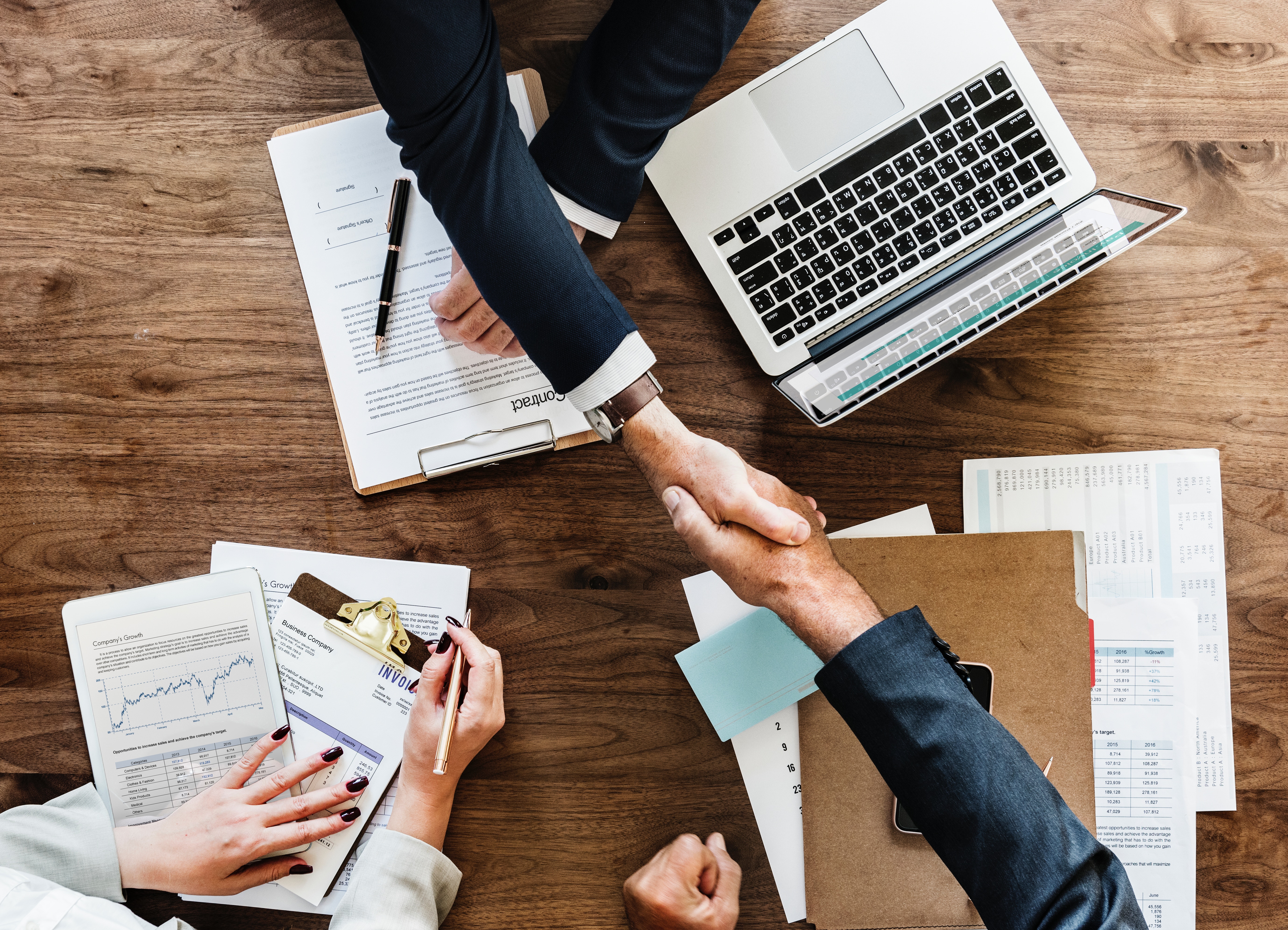 Business handshake over documents with laptop and reports on a wooden table.