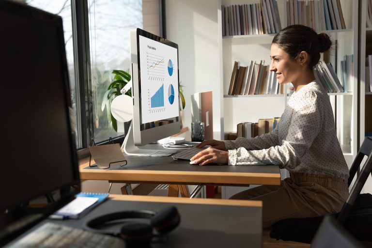 Woman working at a desk with charts and graphs displayed on a computer screen in a home office.
