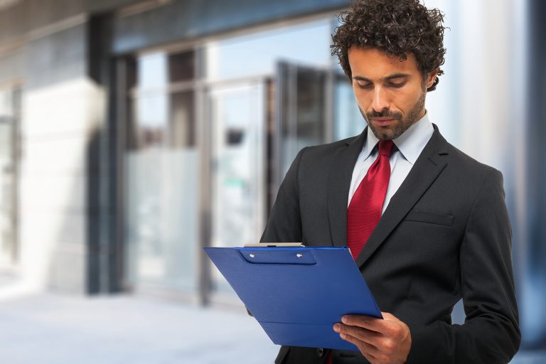 Businessman in a suit reading a document on a clipboard outside an office building.