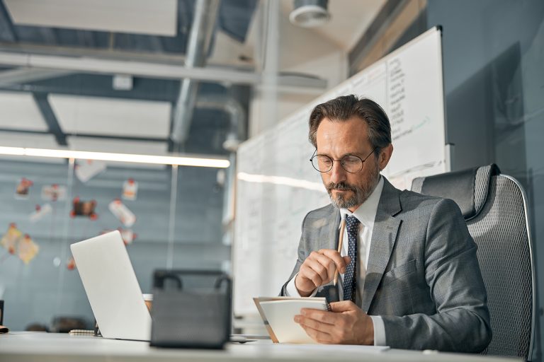 Mature businessman intently using a tablet at his desk in a modern office with a whiteboard in the background.