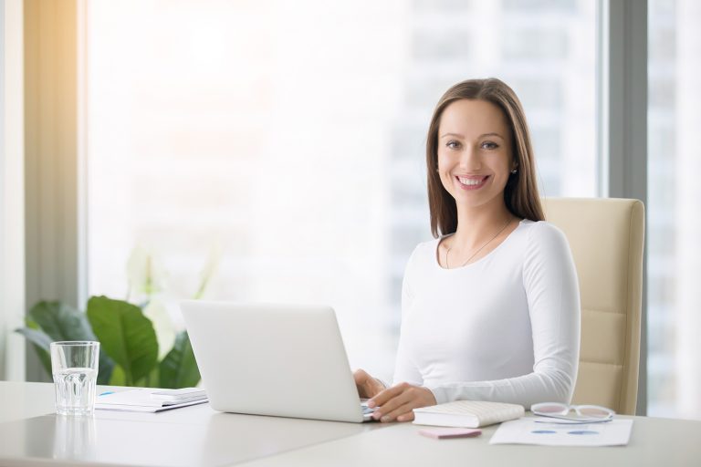 Smiling young woman working on a laptop at a bright office desk with cityscape visible through the window.