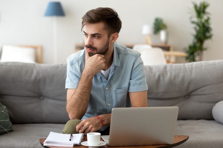 Focused young man with a beard working from a laptop on a couch in a cozy home setting.