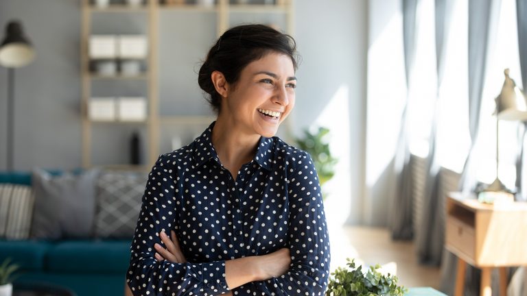 Cheerful woman with crossed arms standing in a well-lit home office, looking away with a joyful expression.