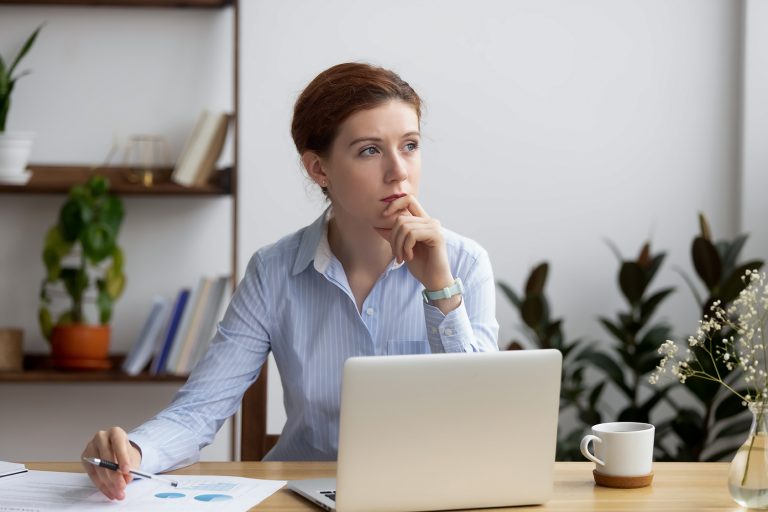 Thoughtful young woman working at a laptop with documents, in a home office with plants and coffee.
