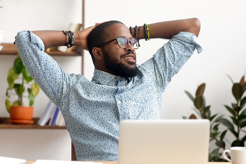 Relaxed professional smiling with hands behind head in front of a laptop.