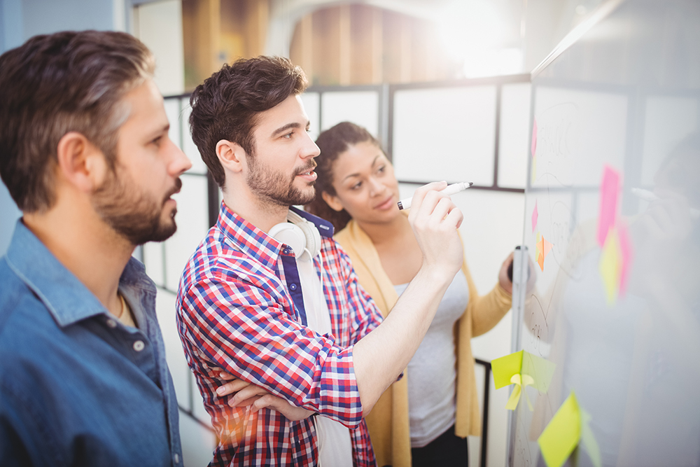 Three professionals collaborating and writing on a glass wall with sticky notes in a creative office.