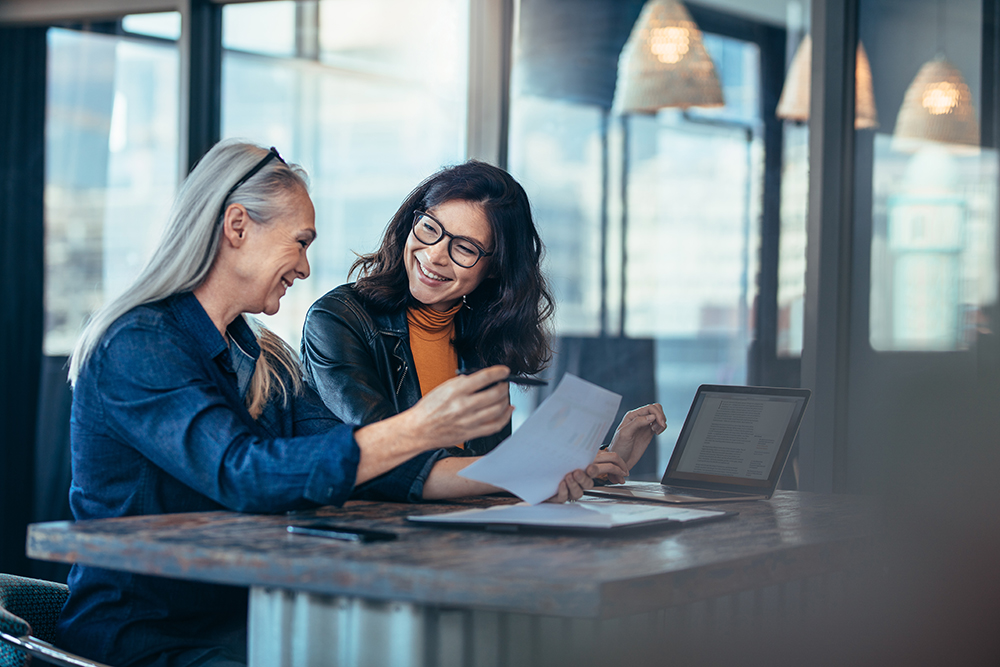 Two professional women smiling and reviewing documents together in a modern workspace.