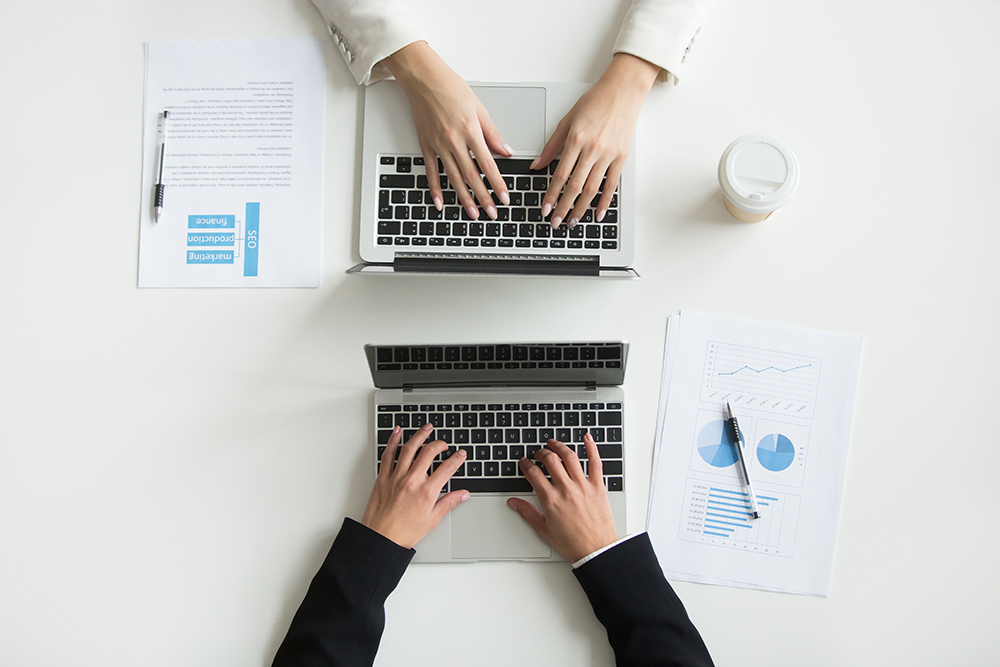 Top view of two professionals typing on laptops with business reports and coffee cup.
