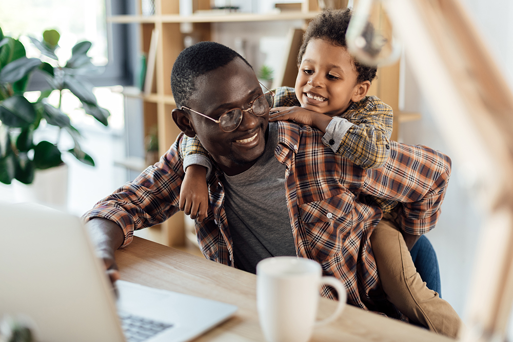 Smiling man with glasses working on laptop with child hugging him from behind at home.