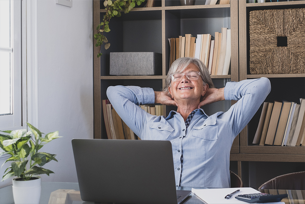 Relaxed senior professional practicing mindfulness at a home office setup with laptop and plants.