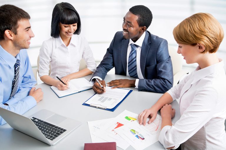 Diverse group of smiling professionals collaborating around a table with laptops and documents.