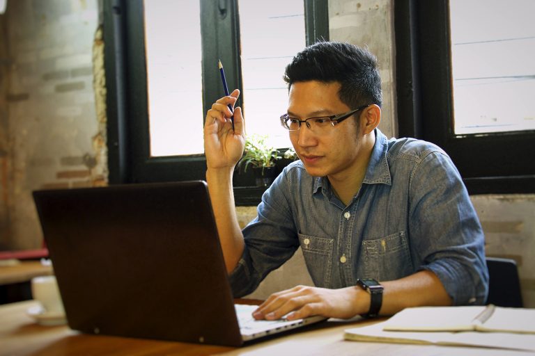 Focused young man working on a laptop at a rustic desk, raising his hand with a pencil.